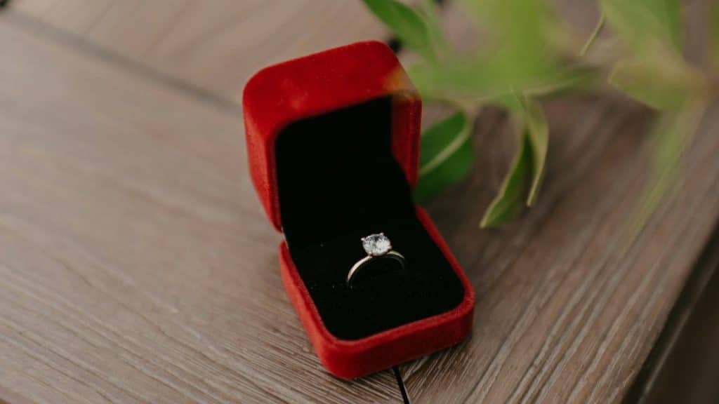 A silver ring with a clear stone sits inside an open, red velvet jewelry box.