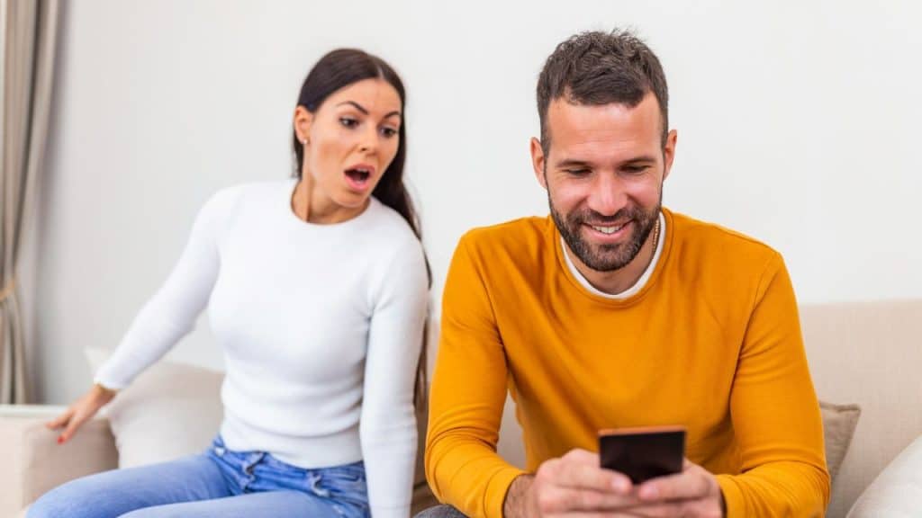 A woman looks surprised while a smiling man in a yellow sweater uses his phone.