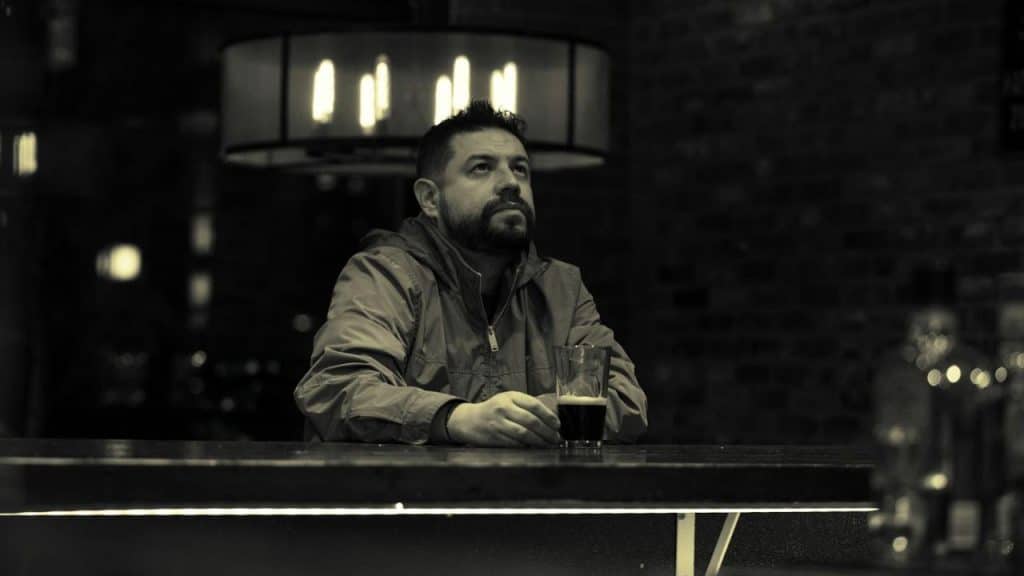 A bearded man in a jacket sits at a dark bar counter holding a drink glass.