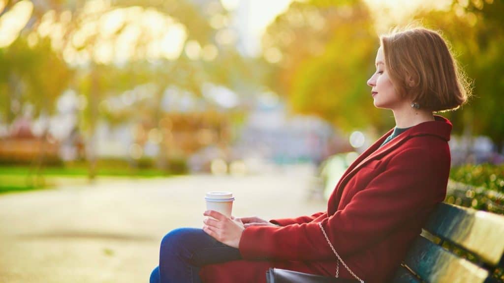 A woman in a red coat sits on a park bench holding a cup.