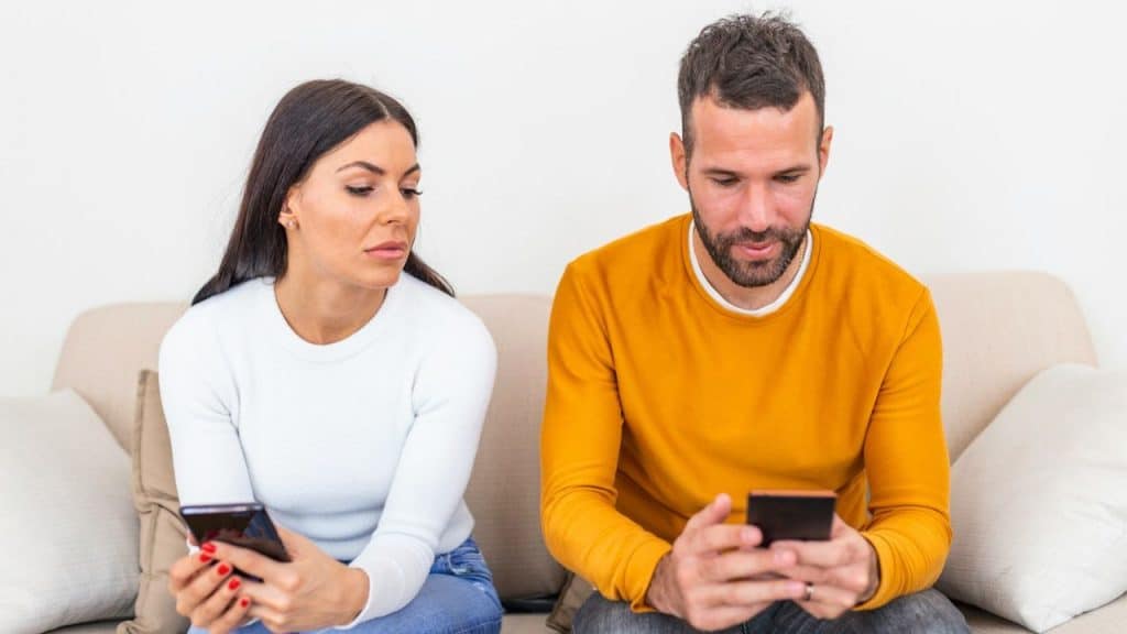 A woman on a couch looks over at a man’s phone while holding her own.