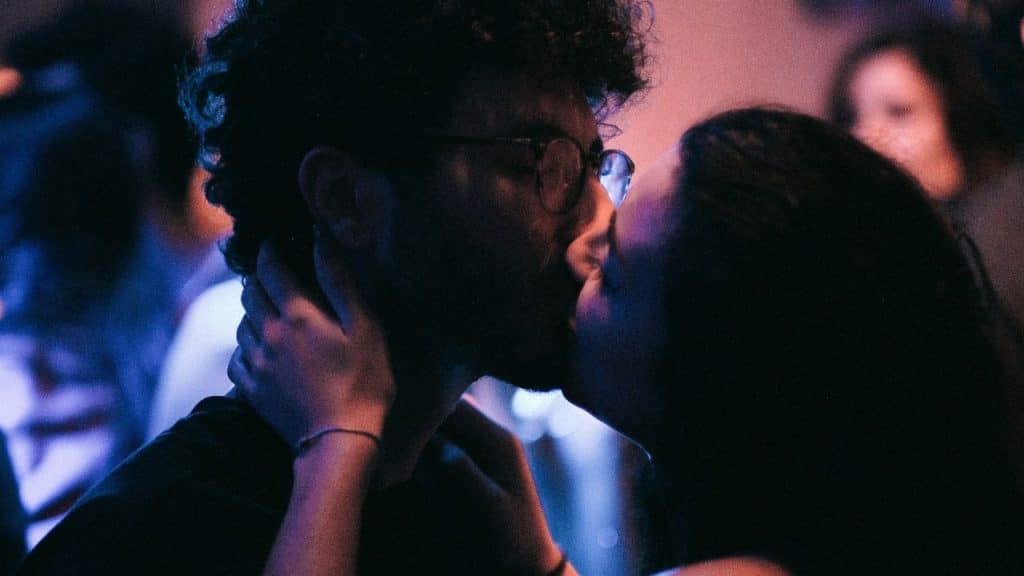 A close-up profile view of two people kissing in a dark room with purple lighting.