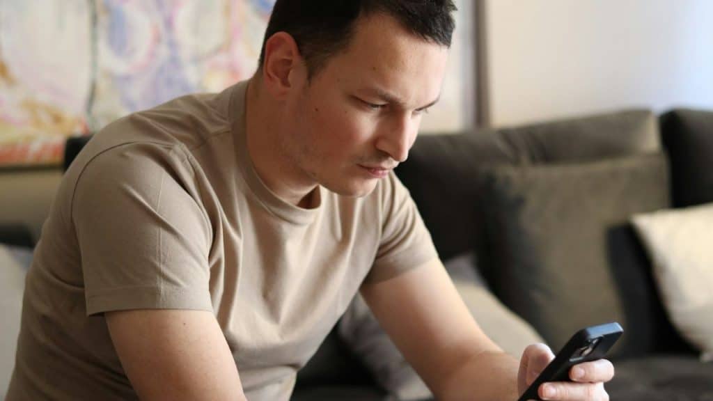 A man in a tan t-shirt sits on a dark sofa while looking down at a smartphone.