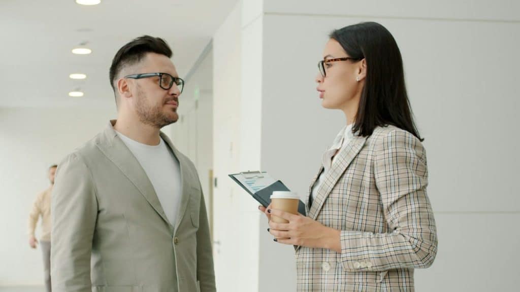 A woman in a blazer holds a clipboard and coffee while speaking to a man.