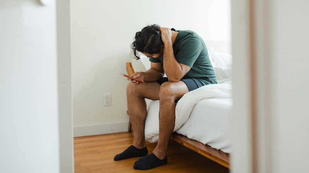 A man sits on the edge of a bed, looking down at his mobile phone.
