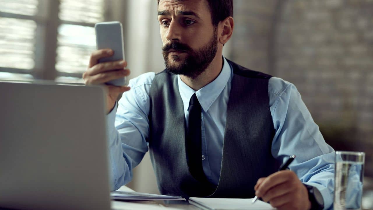 A man with a beard holds a smartphone while writing in a notebook near a laptop.