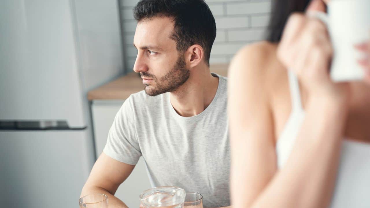 A man in a grey shirt looks sideways while a woman's blurred arm holds a cup.