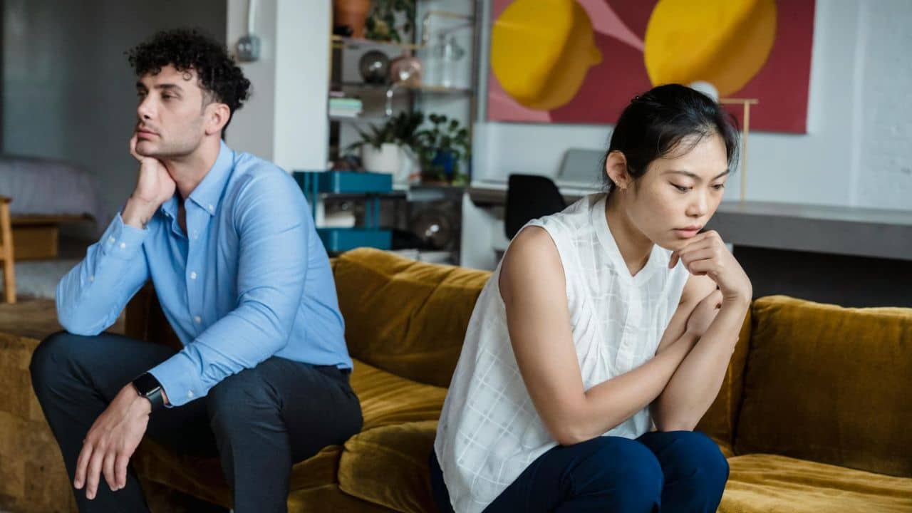 A man and woman sit on a yellow sofa, both looking away with neutral expressions.