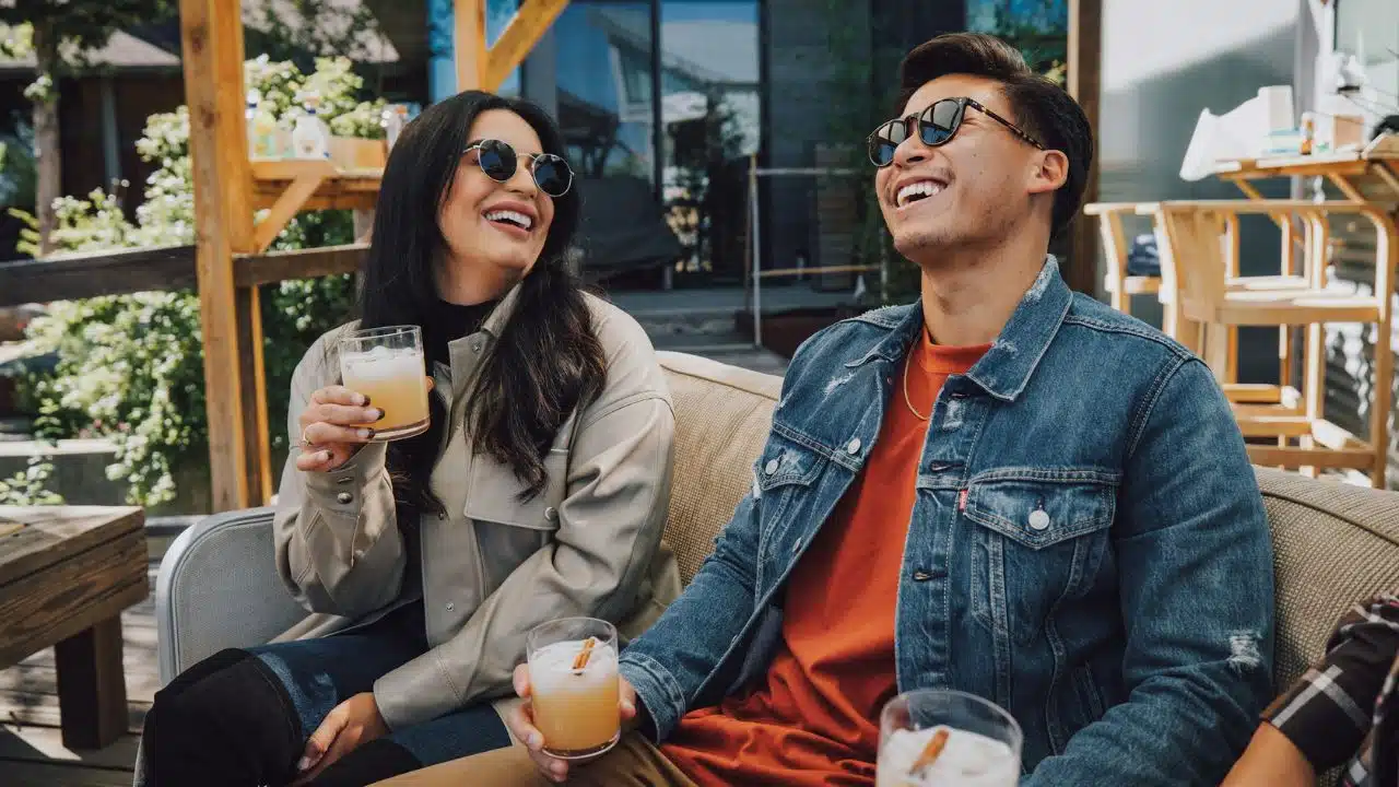 A man and woman laugh while sitting outdoors and holding glasses with drinks.
