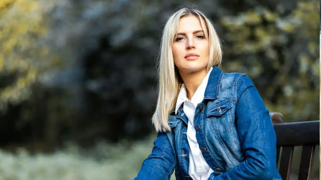 A woman in a blue denim jacket sitting on a brown wooden bench during the daytime.