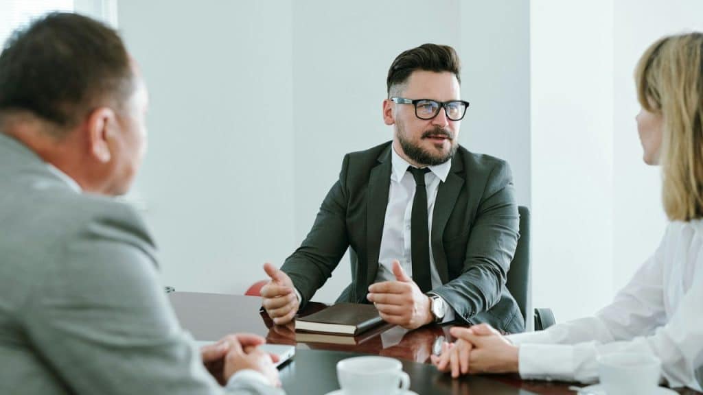 A man in a suit and glasses gestures with his hands during a meeting.