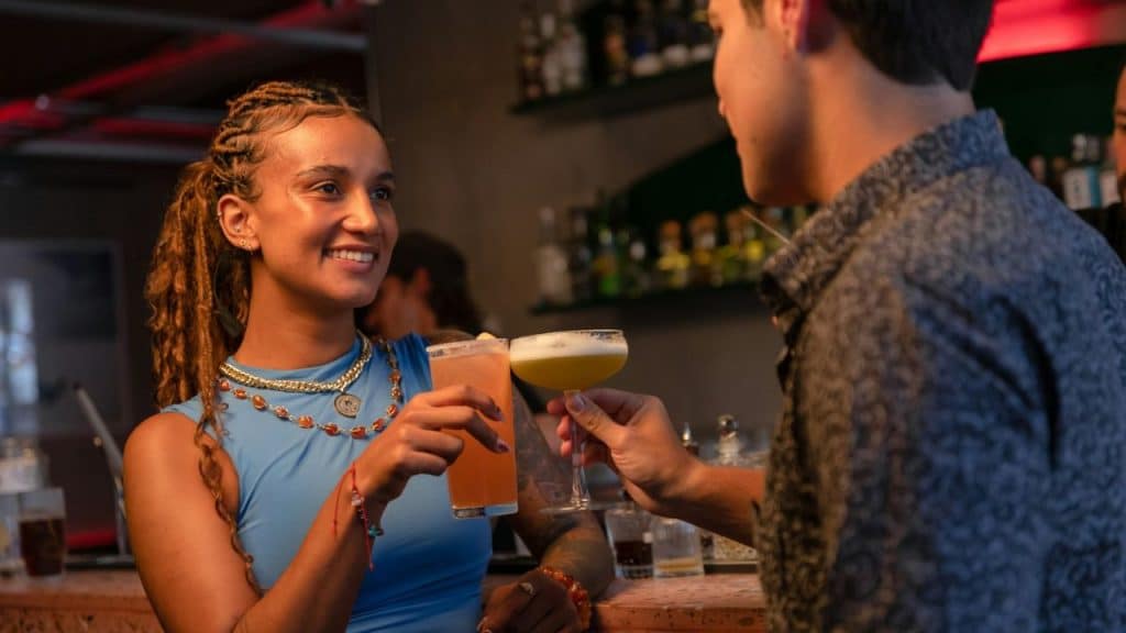 A woman with long braids in a blue top holds a drink and smiles at a man.
