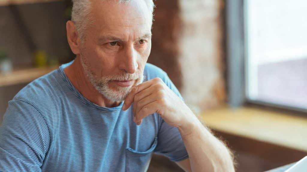 A man with silver hair and a beard looks upward while leaning against a desk.