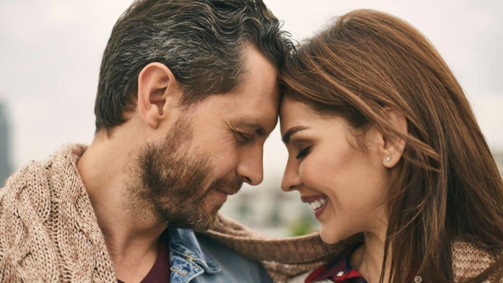 A man and woman with closed eyes lean their foreheads together while smiling softly outdoors.