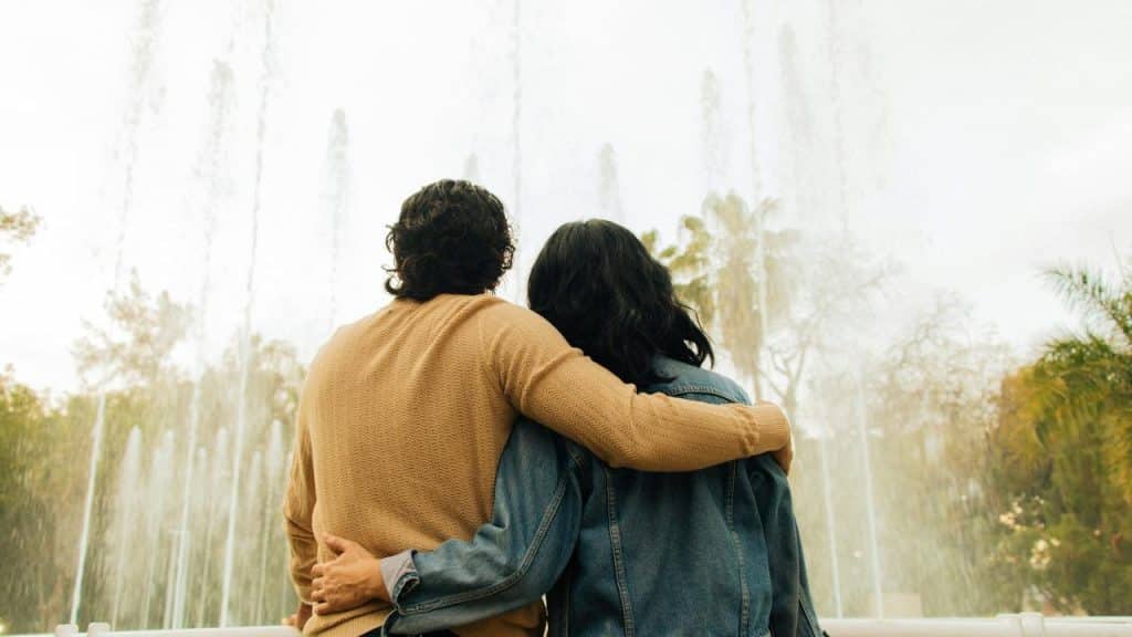 A couple standing arm in arm, viewed from behind, watching a fountain in a park.