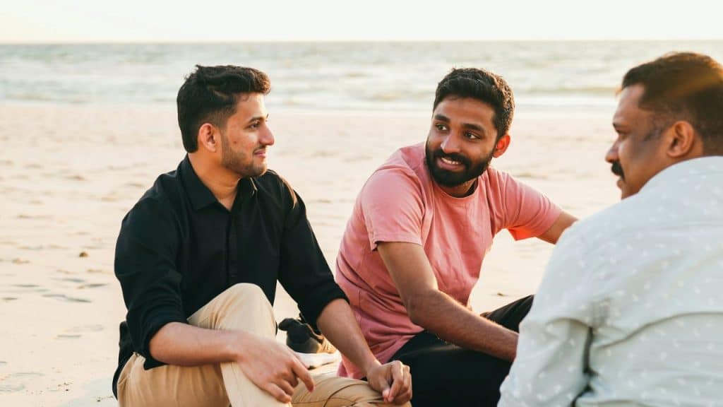 Three men sit on a sandy beach at sunset, engaged in conversation.