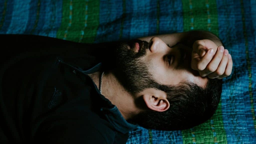 A bearded man lies awake on a blue and green patterned blanket, staring upward.