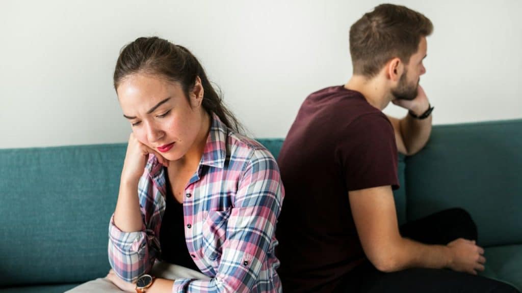 A couple sits back-to-back on a teal couch, looking away from each other in silence.