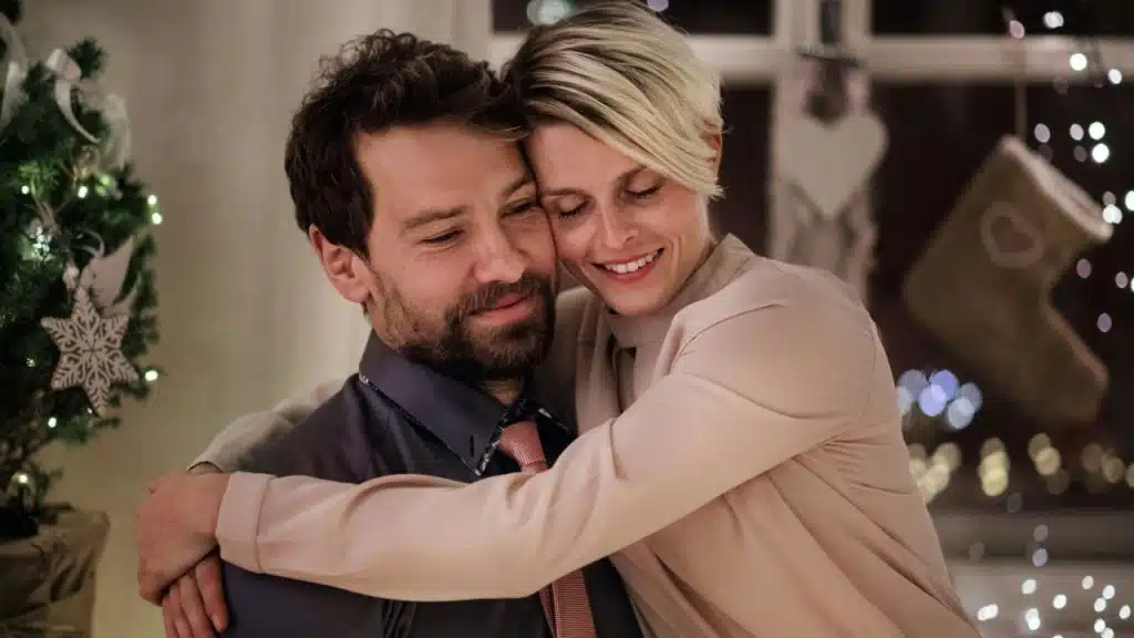 A blonde woman and a bearded man embrace while smiling in a festive, decorated room.