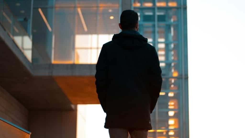 A person in a black hooded jacket stands facing a modern glass building at dusk.
