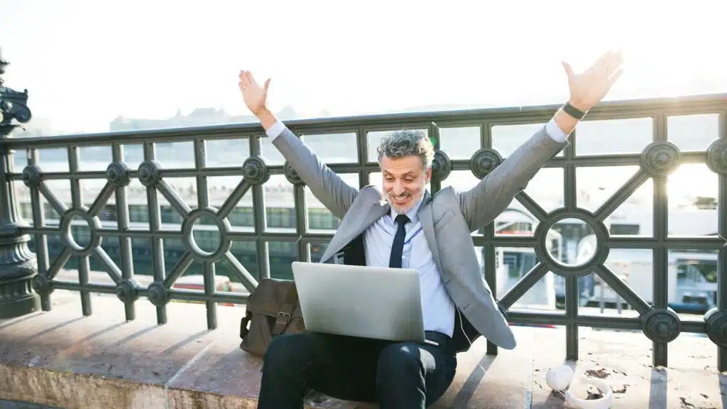 A handsome, mature businessman with a laptop in a city sitting on a bridge stretching his arms.