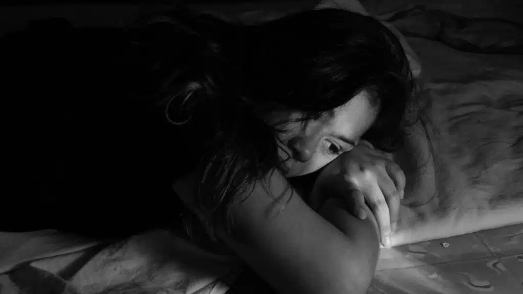 A close-up, black-and-white photo shows a woman lying down with a despondent or weary expression.