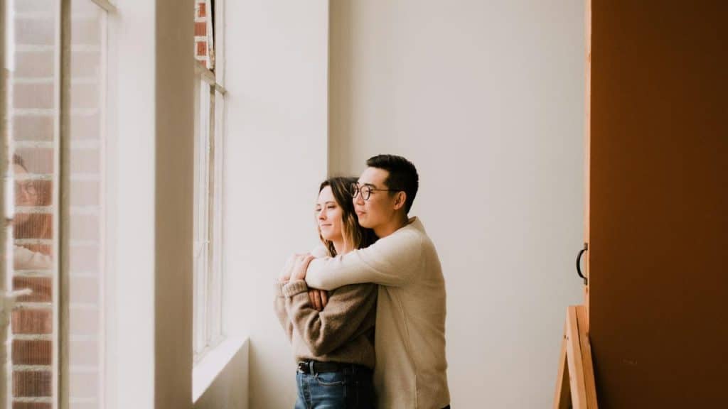 A couple standing by a window while embracing and looking outside.