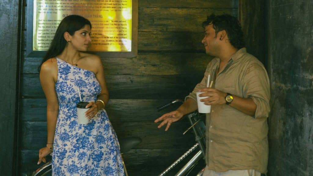 A man and woman standing by a bicycle and talking while holding drinks in a dimly lit setting with a red lantern.
