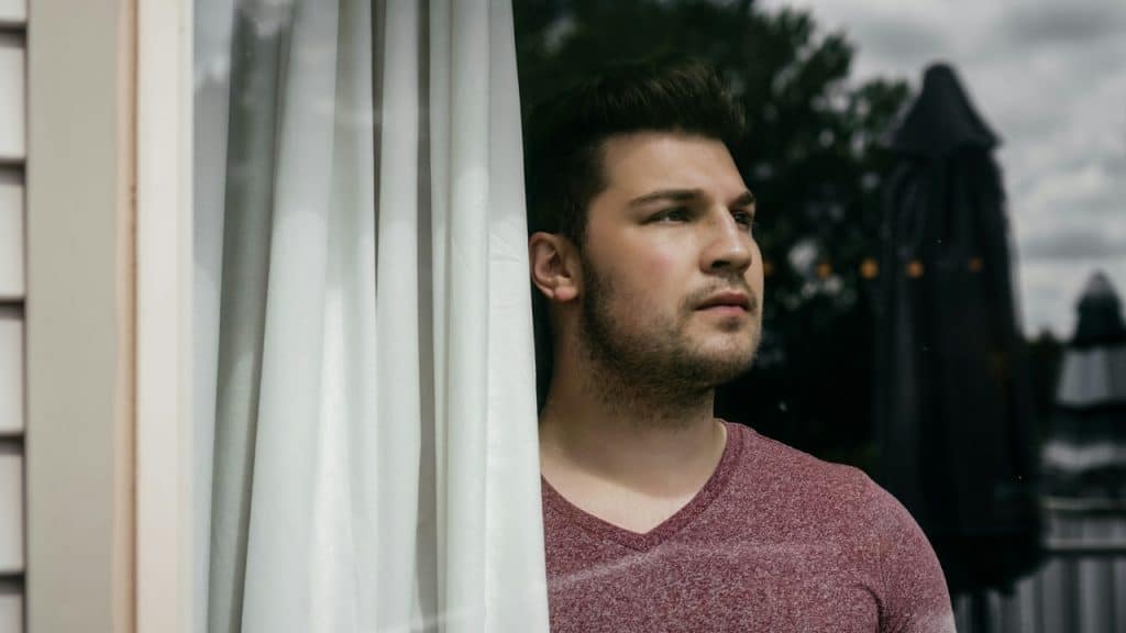 A man with a beard looks out a window next to a white curtain.