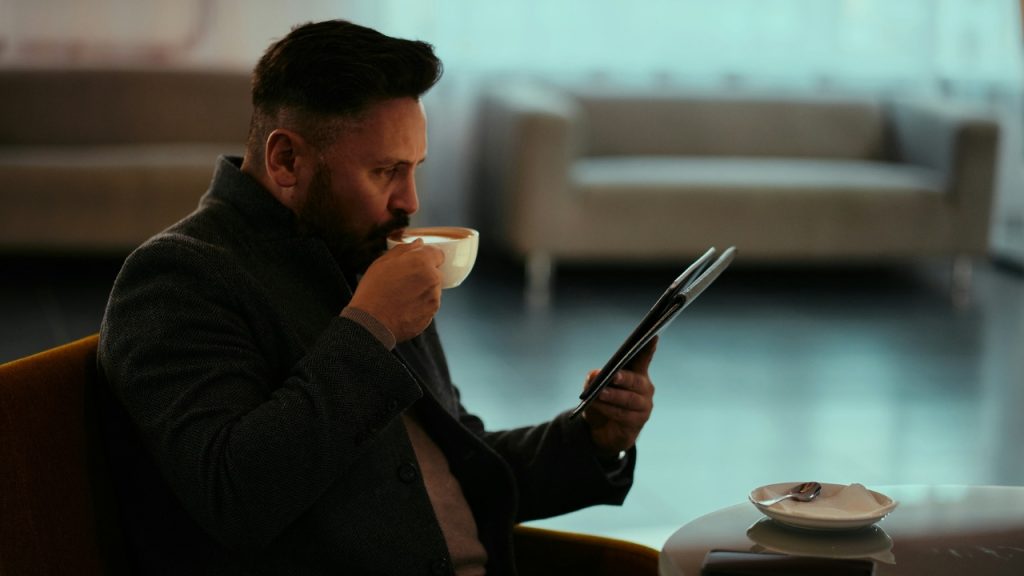 A man in a dark coat drinks from a cup while looking at a tablet.