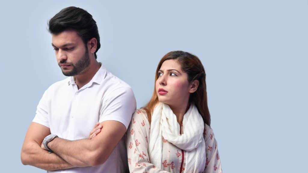 A man with crossed arms looks down while a woman looks at him from behind.