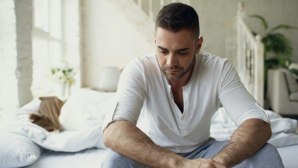 A sad man sitting on the edge of the bed looking down while a woman lies asleep or turned away behind him.