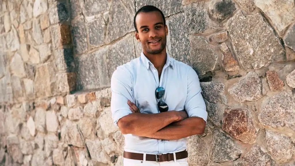 A handsome man in smart casual clothes keeping arms crossed and looking at camera with  smile while standing against the stoned wall outdoors.