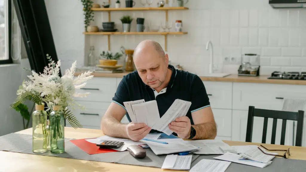 A bald man in a dark polo shirt reviews several papers at a kitchen table.