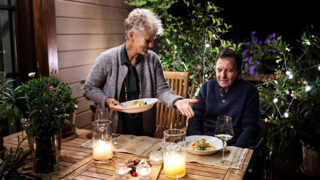 A smiling woman serves food to a man at a candlelit outdoor dinner table.