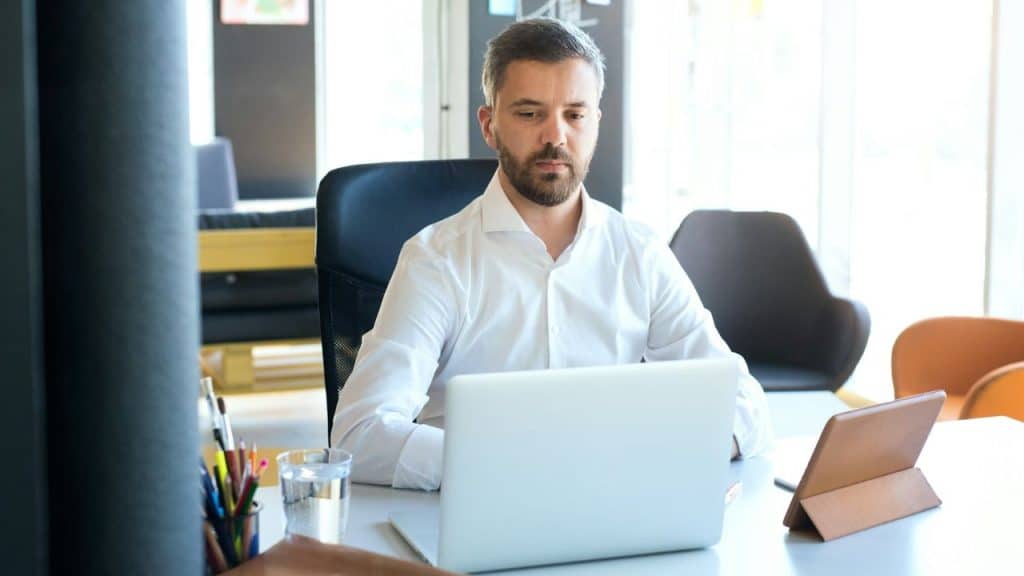 A bearded man in a white dress shirt works on a laptop at an office desk.