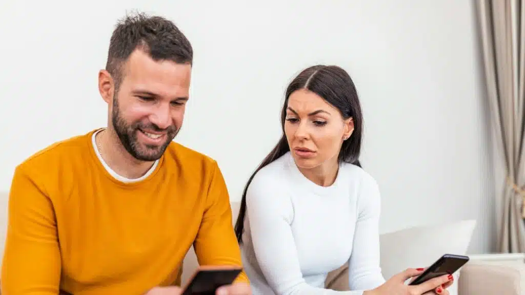 A man smiles at his phone while a woman looks at him with a concerned expression.