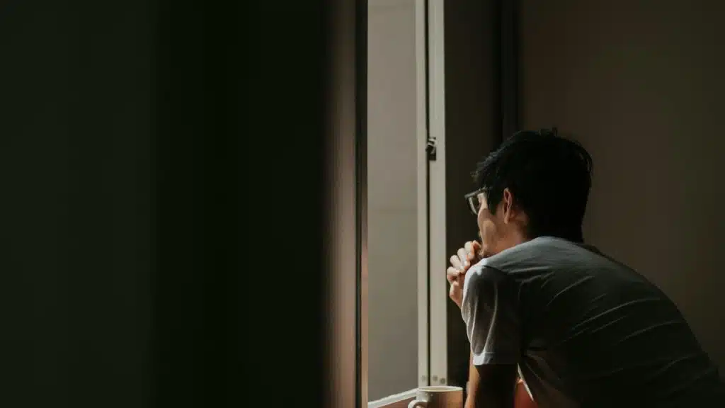 A person wearing glasses looks out a window while holding a mug in soft indoor light.