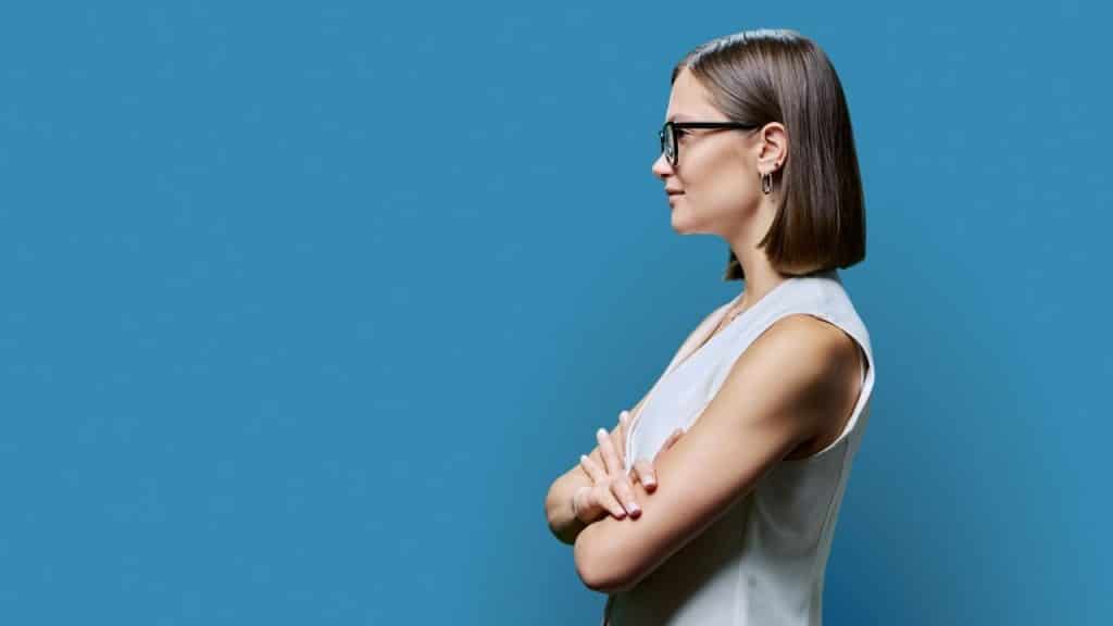 A woman with glasses stands with her arms crossed against a solid blue background.