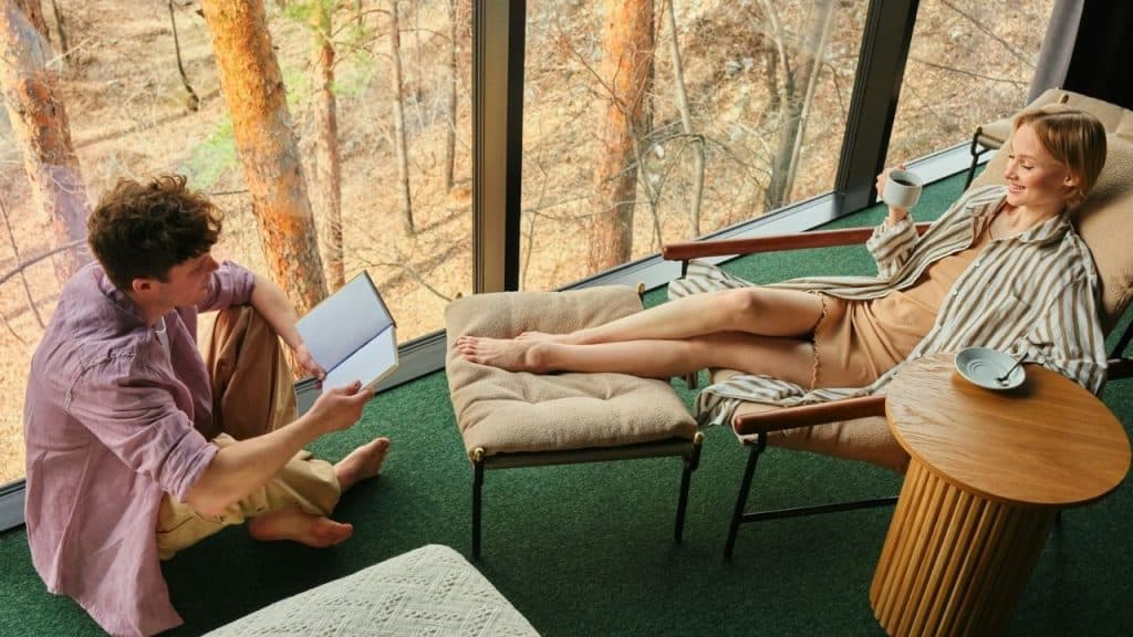 A man reads a book on the floor while a woman relaxes nearby with coffee.