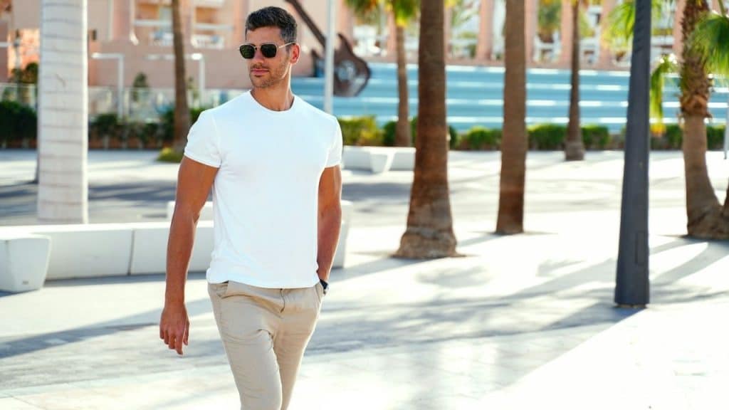 A man in a white t-shirt and sunglasses walks confidently through a sunlit outdoor plaza.