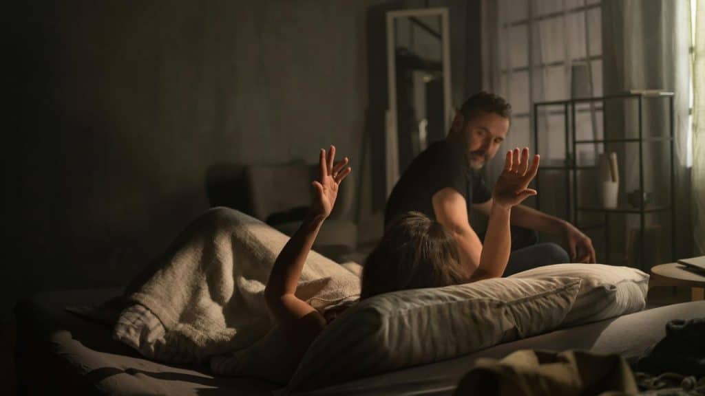 A man sitting beside a bed as a person lies under a blanket with raised hands.