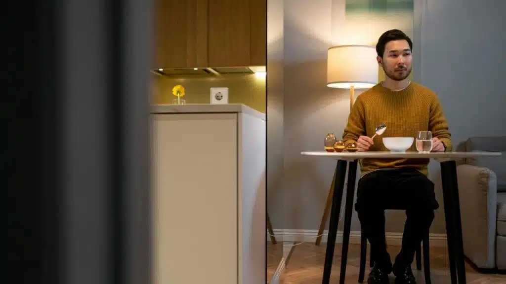 A man in a yellow sweater sits alone at a small table eating with a spoon.
