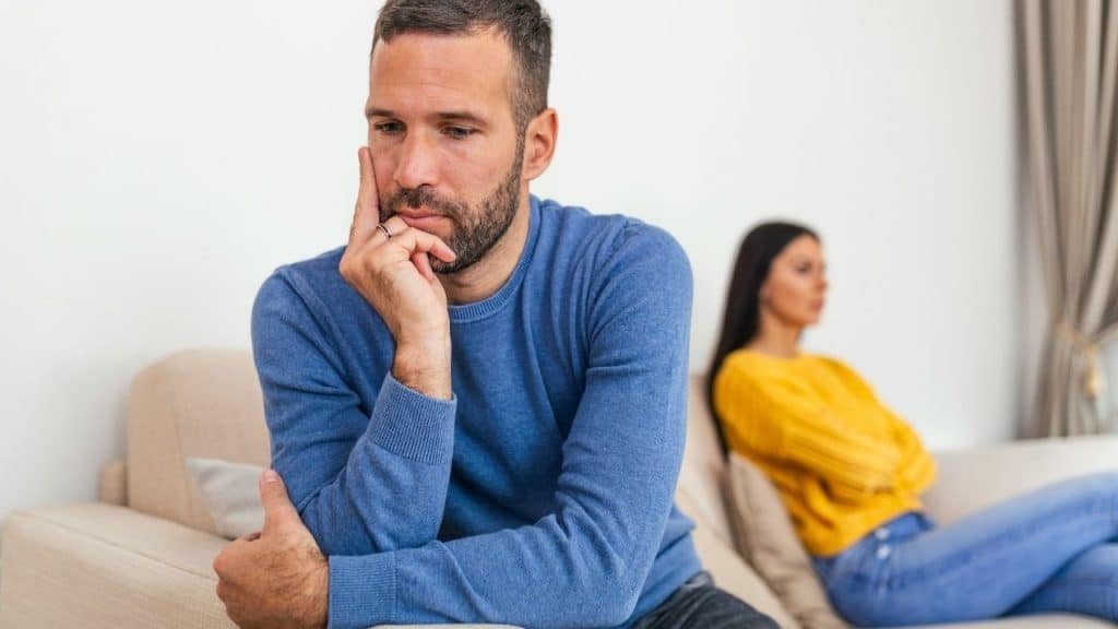 A man in a blue sweater sits on a couch looking pensive while a woman sits behind him.