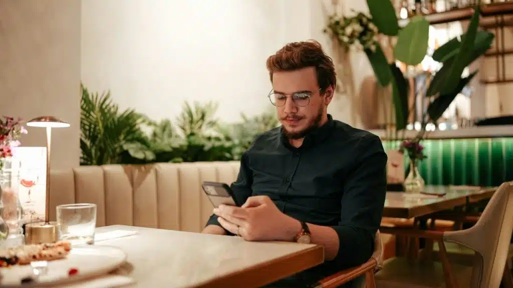 A man wearing glasses sits alone at a restaurant table while looking at his phone.