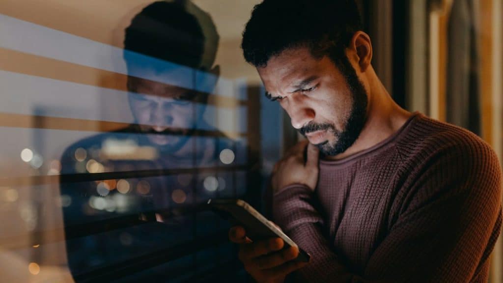 A man with a beard looks down at his phone near a window at night.