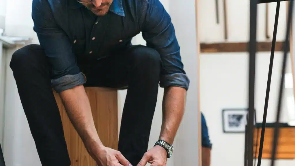 A man crouching indoors, tying his shoes while wearing a denim shirt.