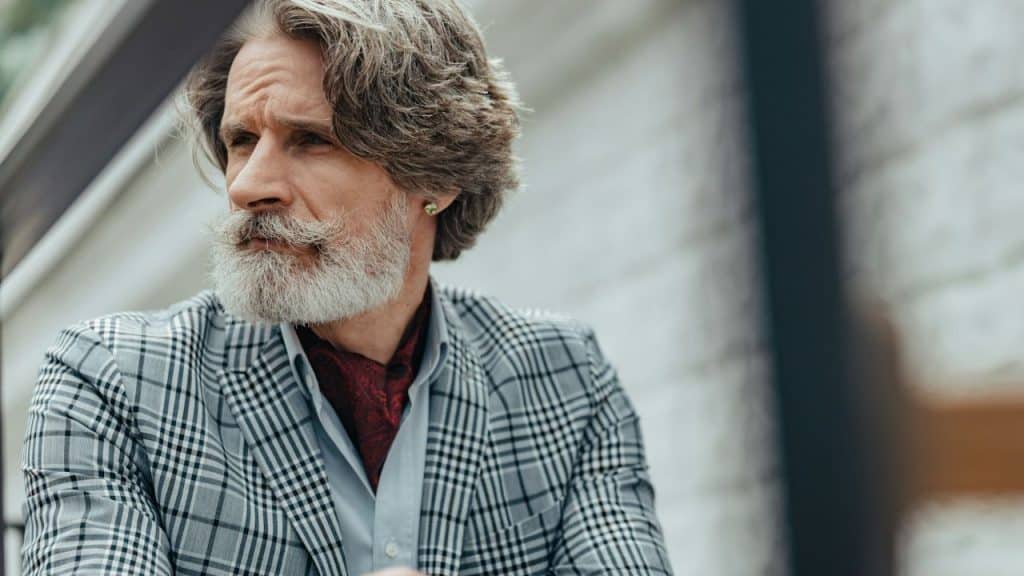 A man with a grey beard and patterned blazer looks thoughtfully into the distance.