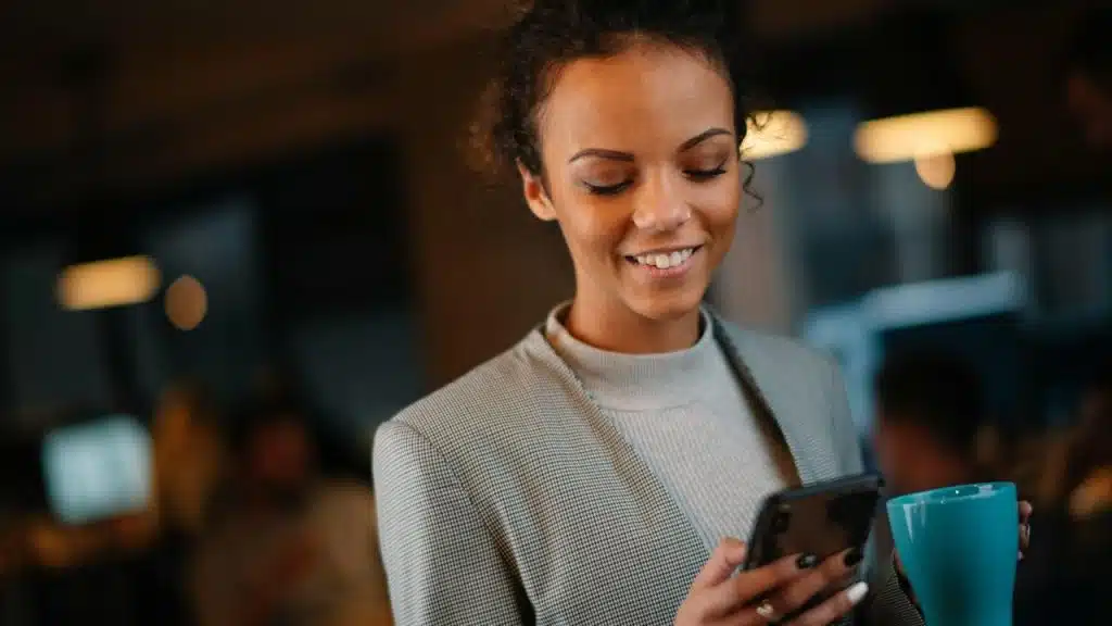 A woman smiling while checking her phone and holding a coffee cup.