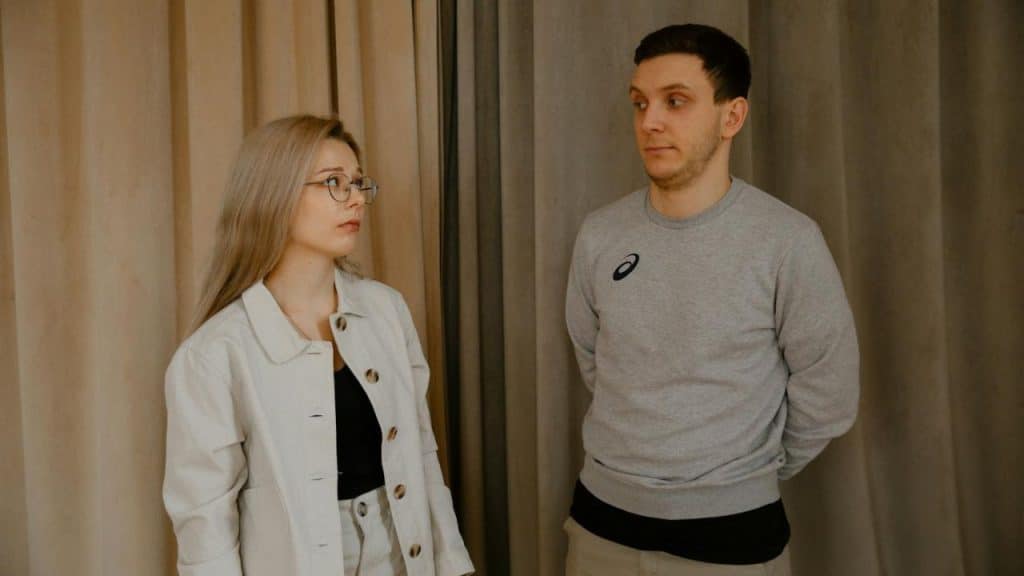 A woman with glasses and a man in a gray sweatshirt stand before beige curtains.